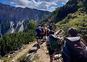 Eine Gruppe Kinder und Erwachsener mit Wanderrucksächen gehen hintereinander über einen Bergpfad