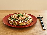 Teller mit einem mit Saucen und Kräutern schön angerichteter japanischer Pfannkuchen, neben dem Teller liegt ein paar Eßstäbchen