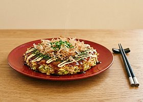 Teller mit einem mit Saucen und Kräutern schön angerichteter japanischer Pfannkuchen, neben dem Teller liegt ein paar Eßstäbchen