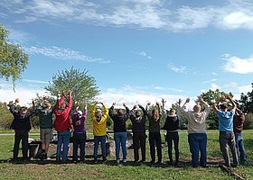 10 junge Menschen mit hochgestreckten Armen auf einem Feld stehend, von hinten fotografiert