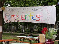 Stoffbanner zwischen Bäumen gespannt, in bunten Buchstaben steht dort "Familienfest", im Vordergrund Tische und Bänke