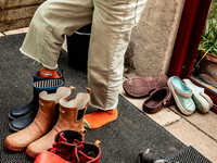 Man sieht die Beine eines Kindes, in beiger Hose und orangenen Strümpfen. Das Kind schlüpft in ein paar Gummistiefel, die unter vielen anderen großen und kleinen Schuhen im Eingangsbereich eines Hauses stehen.