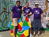 zwei Mitarbeiter und eine Mitarbeiterin in lila T-Shirts mit Logo der Kinderspielstadt vor Rapunzelturm-Kulisse