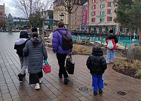 Kinder und ein Betreuer mit Sporttaschen und Rücksäcken, von hinten fotografiert, im Hintergrund ein Außenschwimmbecken