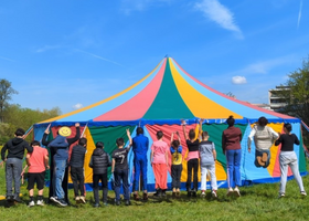 eine Reihe von Kindern springt vor einem bunten Zirkuszelt in die Luft, sie sind von hinten zu sehen