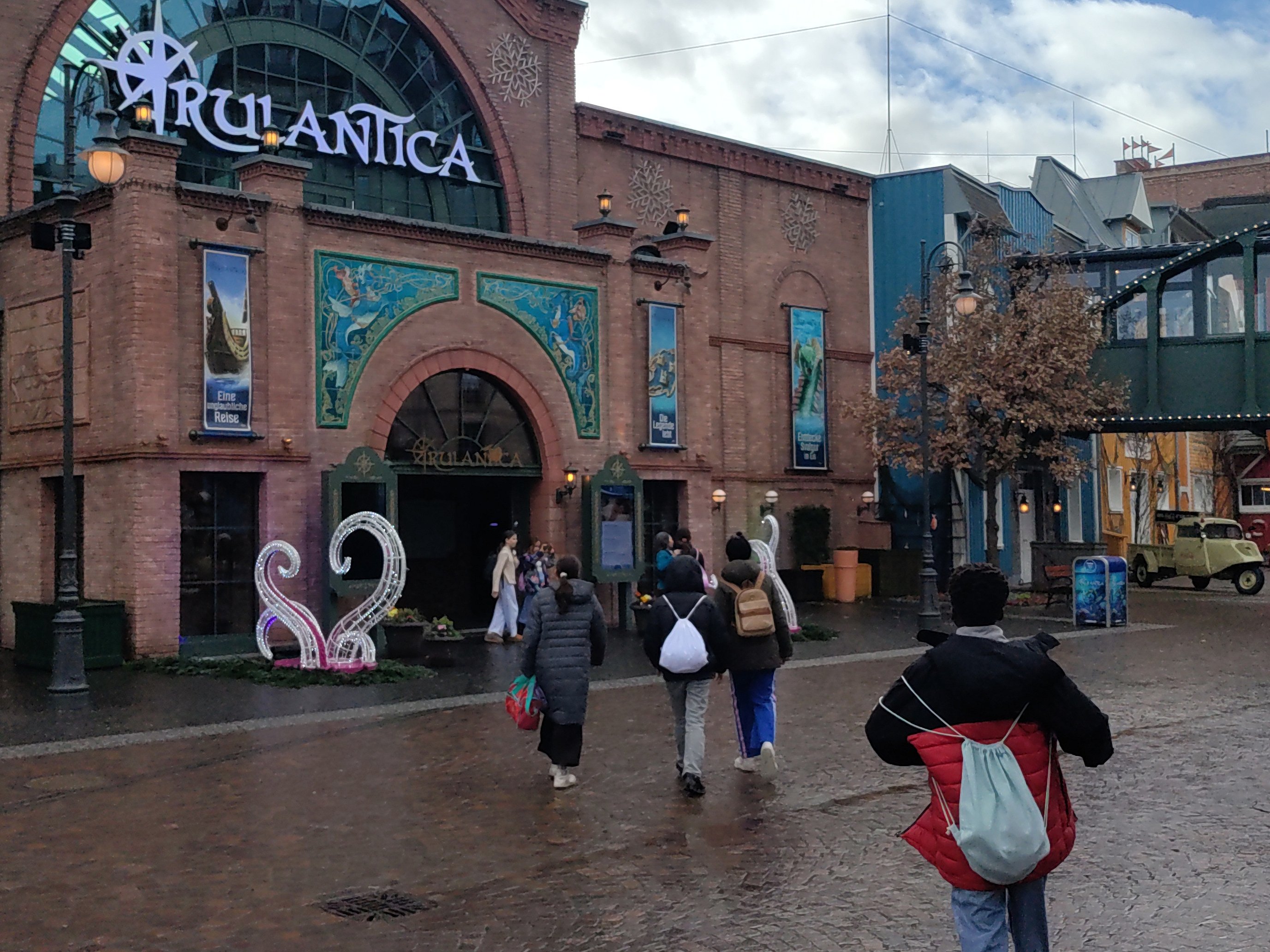 Kinder mit Sporttaschen und Rücksäcken, von hinten fotografiert; sie gehen auf den Eingangsbereich eines Erlebnisbades zu