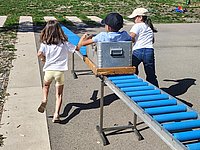 Zwei Kinder führen ein Kind, das in einer Kiste auf einer Rollenrutsche sitzt