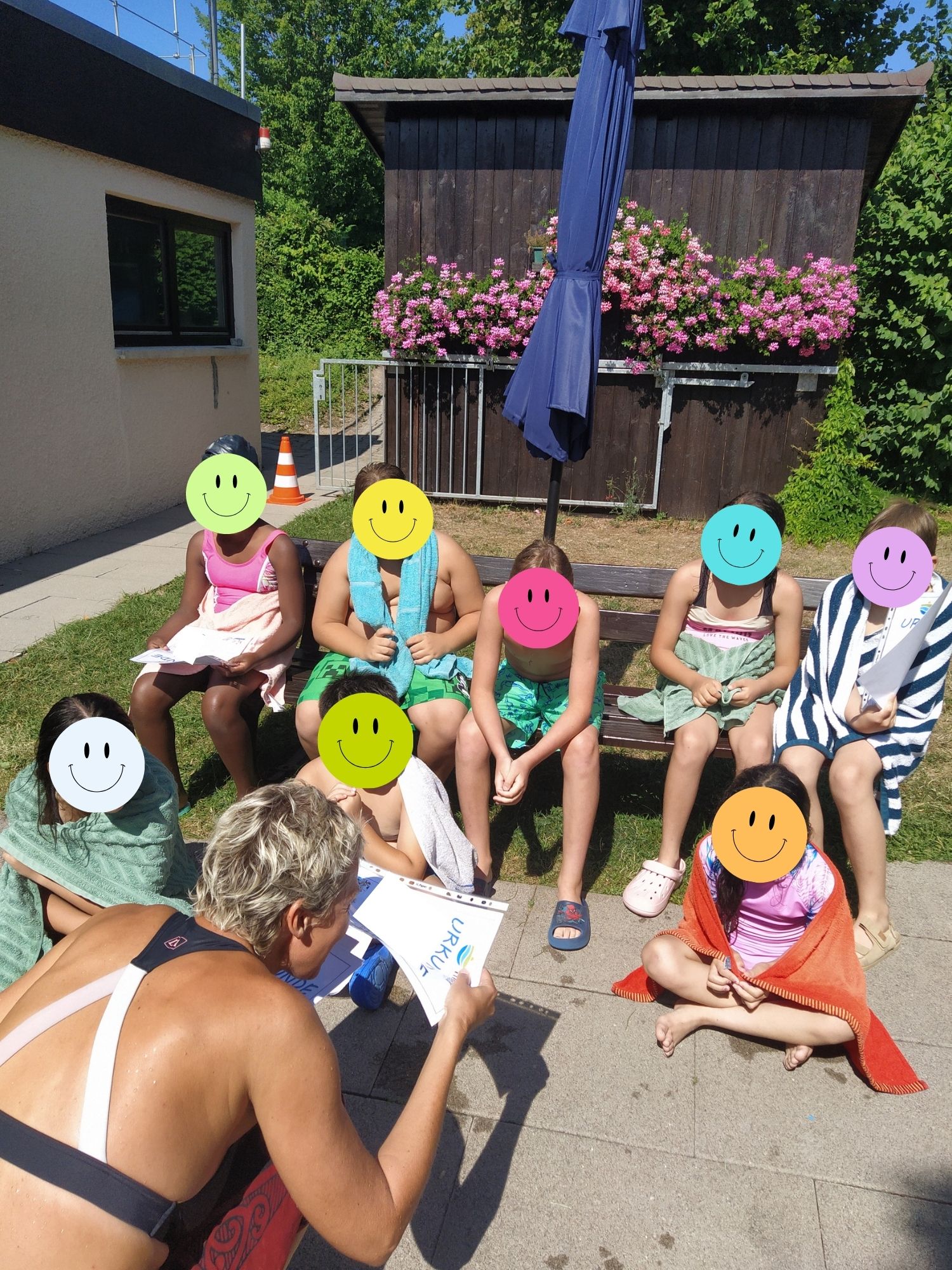 Kinder in Badebekleidung, unkenntlich durch Smiley auf dem Gesicht, bekommen eine Schwimmurkunde überreicht