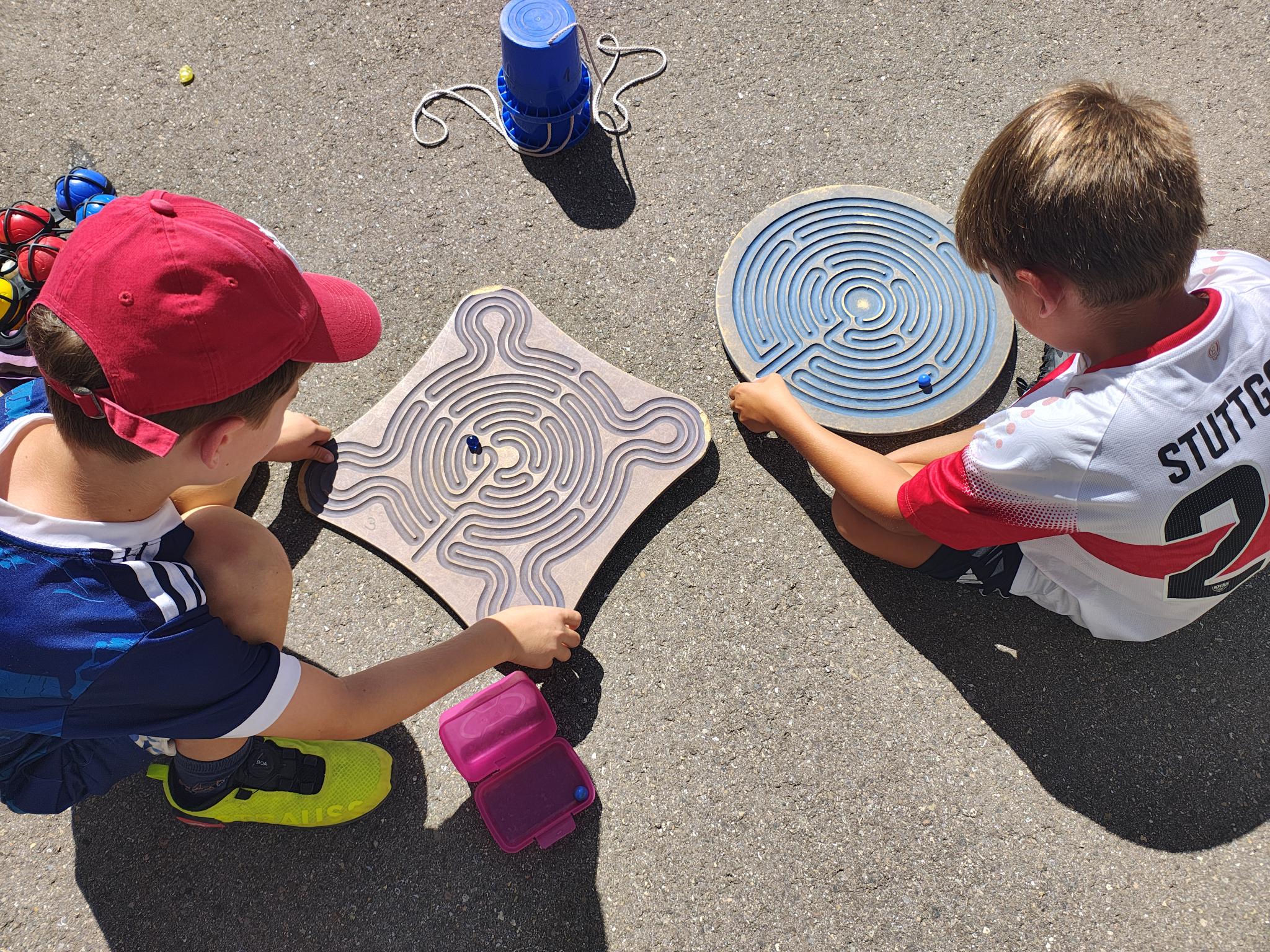 Zwei Kinder (von oben fotografiert) sitzen auf Asphalt und spielen mit  Murmel-Balace-Scheiben